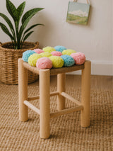 A wooden stool with a natural finish features a colorful top adorned with pink, blue, and yellow pom poms. The stool has four sturdy legs and is placed on a textured beige rug, with a woven basket and a small piece of artwork visible in the background