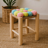 A wooden stool with a light natural finish, featuring a colorful top adorned with pink, blue, and yellow pom poms. The stool has four sturdy legs and is placed on a textured natural fiber rug, with a woven basket and a plant visible in the background