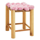 A wooden stool with four legs and a woven seat adorned with fluffy pink pom poms, creating a playful and vibrant look suitable for home decor