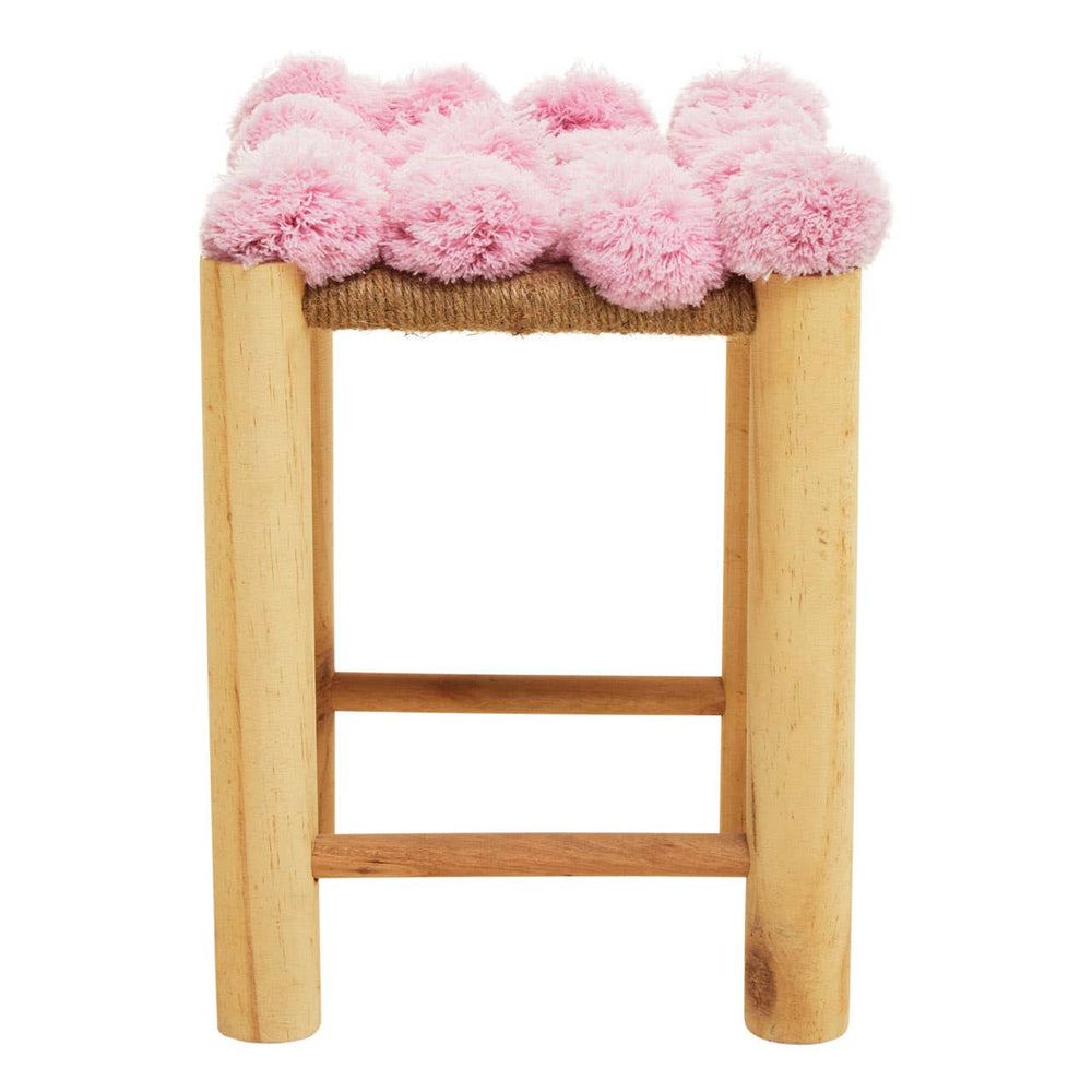 A wooden stool with a natural finish featuring a top covered in soft pink pom poms, creating a playful and whimsical design. The stool has a simple, sturdy structure with a horizontal support bar between the legs