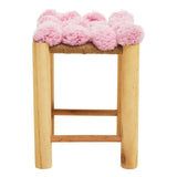 A wooden stool with a natural finish featuring a top covered in soft pink pom poms, creating a playful and whimsical design. The stool has a simple, sturdy structure with a horizontal support bar between the legs