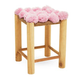 A wooden stool with light-colored legs and a top adorned with fluffy pink pom poms, creating a playful and whimsical design