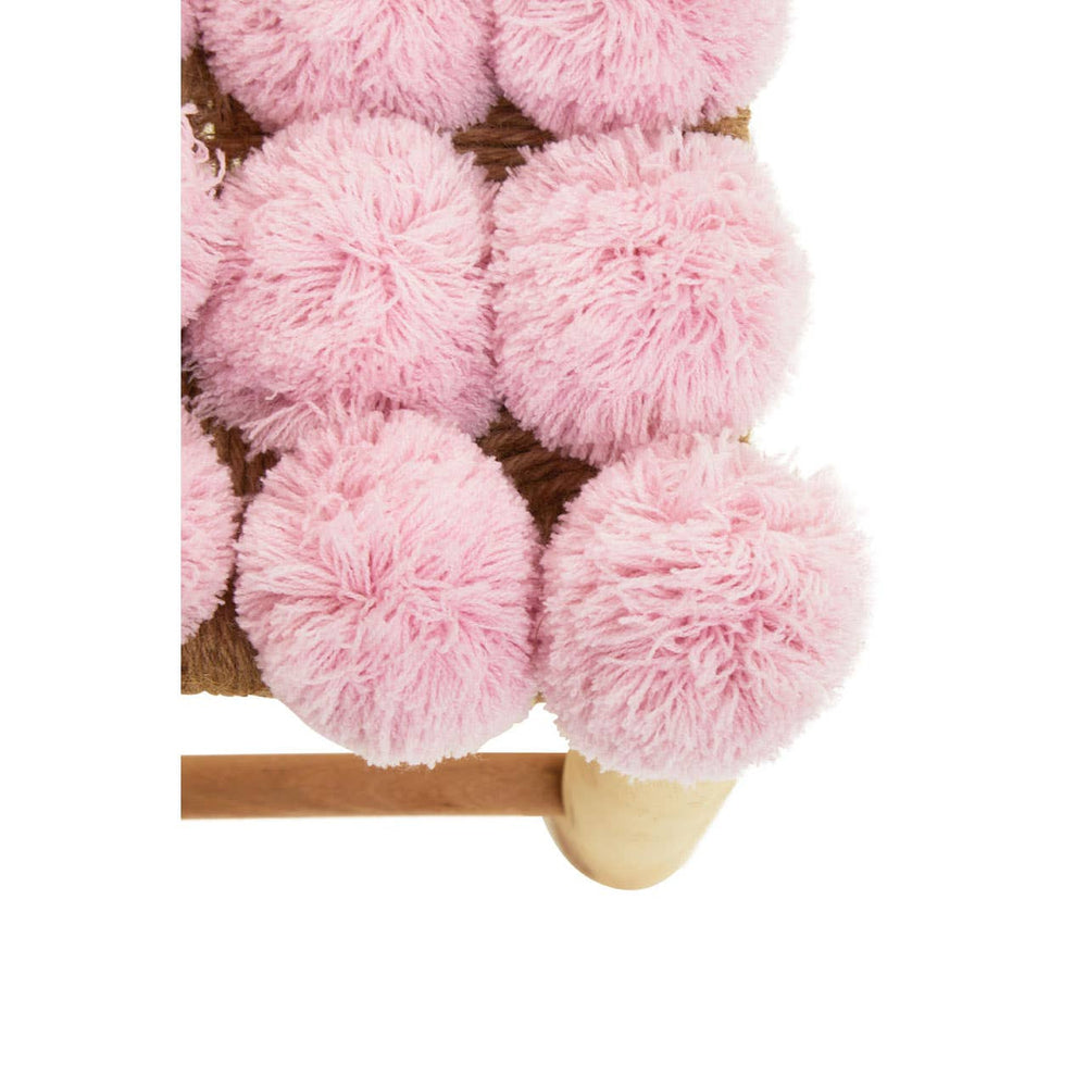 A close-up view of a pink pom pom stool featuring soft, fluffy pom poms arranged in a grid pattern on the seat, with a light wooden base and legs. The pom poms are a pastel pink color, adding a playful and cozy touch to the design