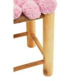 A close-up view of a West Village Pink Pom Pom Stool featuring a soft pink pom pom cushion on top and light wooden legs. The legs are sturdy and rounded, showcasing a natural wood finish