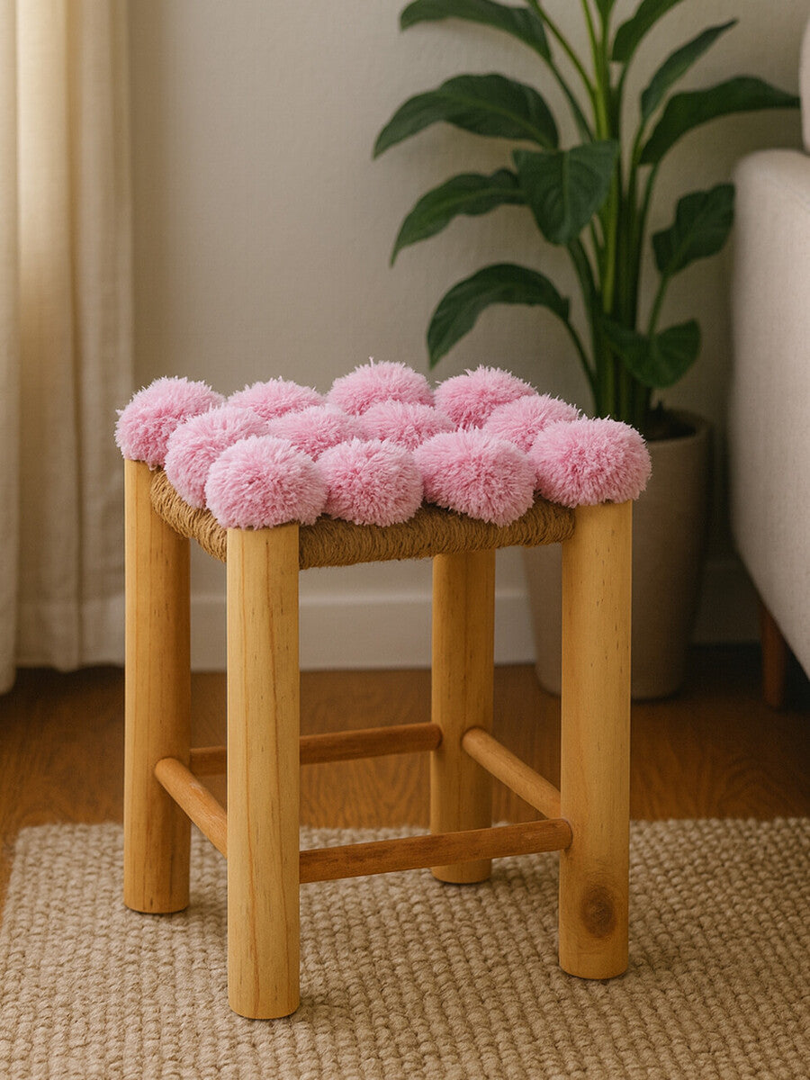 A wooden stool with a natural finish featuring a top adorned with soft pink pom poms. The stool has four sturdy legs and is positioned on a light-colored rug, with a plant visible in the background