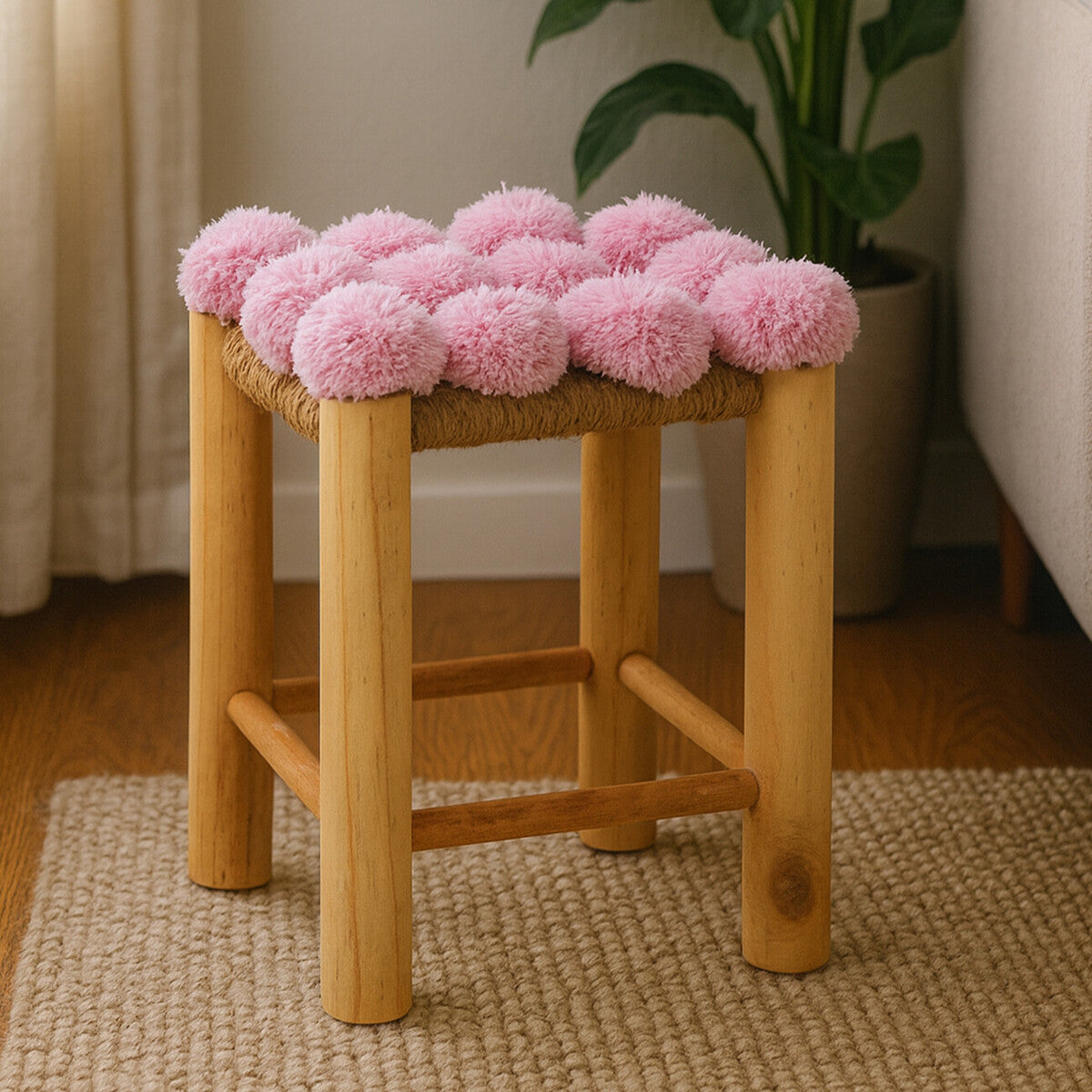 A wooden stool with a natural finish featuring a top covered in soft pink pom poms, placed on a light-colored rug in a cozy room with a plant in the background