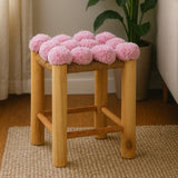 A wooden stool with a natural finish featuring a top covered in soft pink pom poms, placed on a light-colored rug in a cozy room with a plant in the background