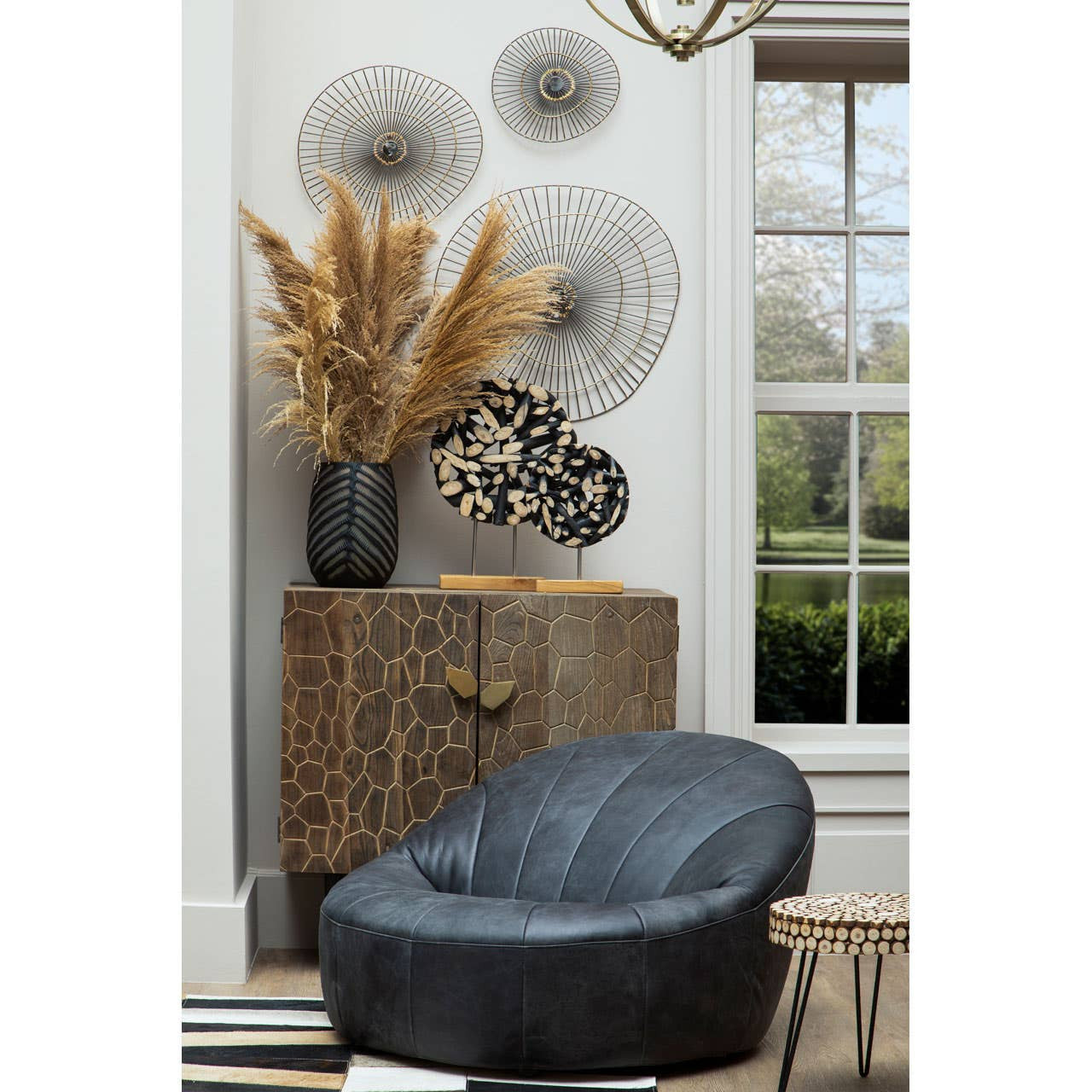 Round side table made from fir wood with a mosaic design, featuring a natural wood finish. The table is positioned next to a black bean bag chair and a textured cabinet. Behind the table, there are decorative wall hangings and a vase with dried flowers, creating a cozy living space ambiance