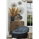 Round side table made from fir wood with a mosaic design, featuring a natural wood finish. The table is positioned next to a black bean bag chair and a textured cabinet. Behind the table, there are decorative wall hangings and a vase with dried flowers, creating a cozy living space ambiance