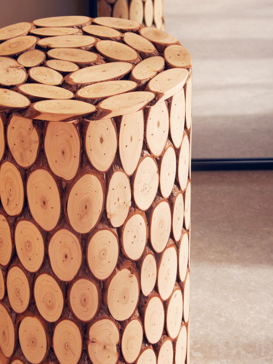 Round side table or stool made from fir wood, featuring a mosaic design of circular wood slices arranged closely together. The surface is smooth and polished, showcasing the natural wood grain and warm tones. The table is positioned on a light-colored floor, with a soft, neutral background