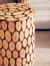 Round side table or stool made from fir wood, featuring a mosaic design of circular wood slices arranged closely together. The surface is smooth and polished, showcasing the natural wood grain and warm tones. The table is positioned on a light-colored floor, with a soft, neutral background