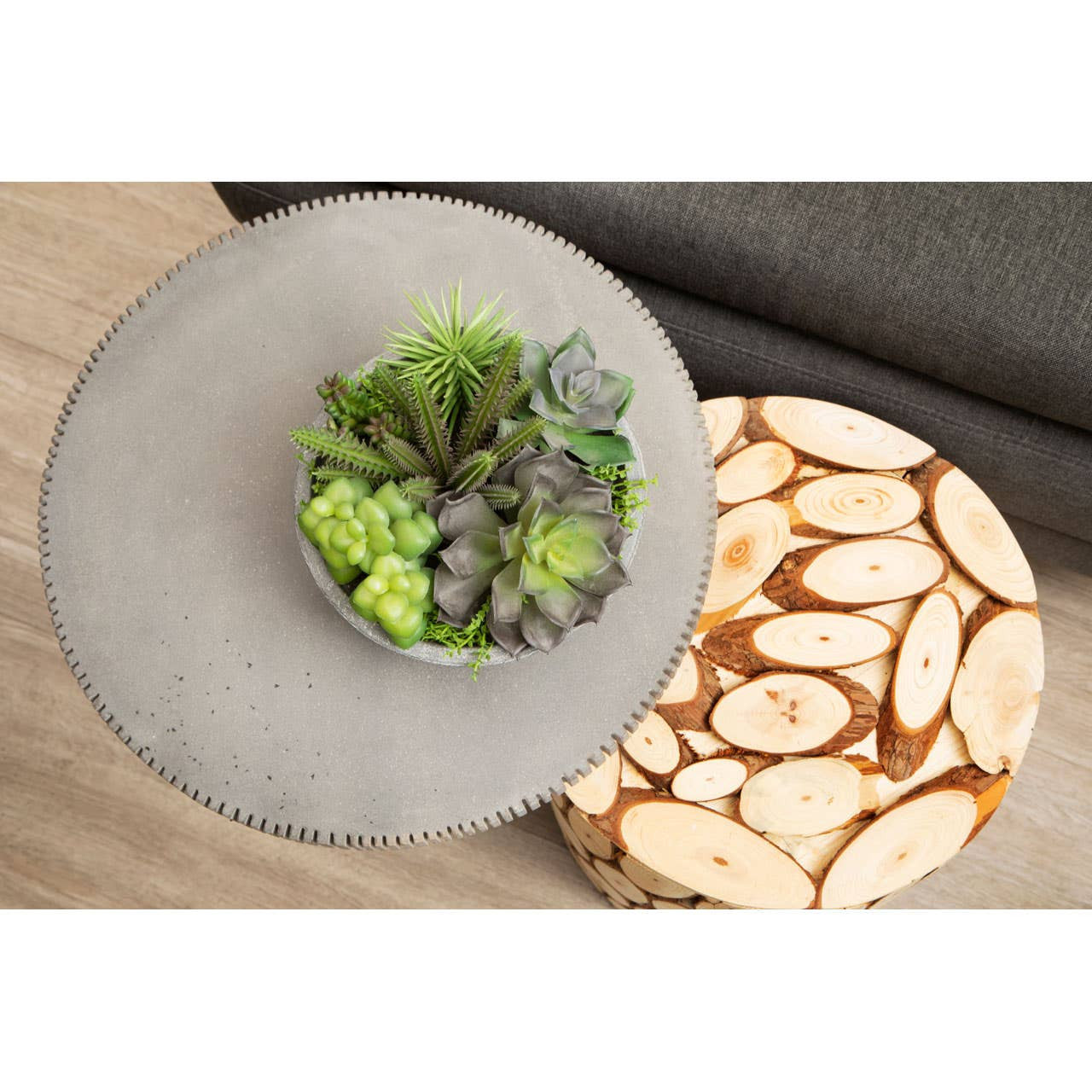 A round side table with a smooth gray top, featuring a small arrangement of green succulents in a decorative pot. The base is made of fir wood with a mosaic design, showcasing natural wood slices in varying shades of brown and cream. The table is placed on a light-colored surface, with a dark fabric visible in the background
