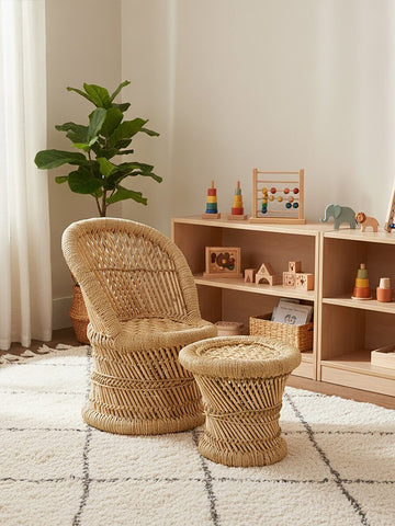 Natural woven bamboo chair and stool set designed for children, featuring a curved chair with a comfortable backrest and a matching round stool. The furniture is placed in a bright, airy room with a light-colored rug and wooden shelving displaying colorful toys and decor. A green plant adds a touch of nature to the space