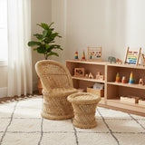 Natural woven bamboo chair and matching stool set designed for children, positioned in a bright room with a light-colored rug and a wooden shelf displaying colorful toys and decor. A green plant is visible in the background, adding a touch of nature to the space