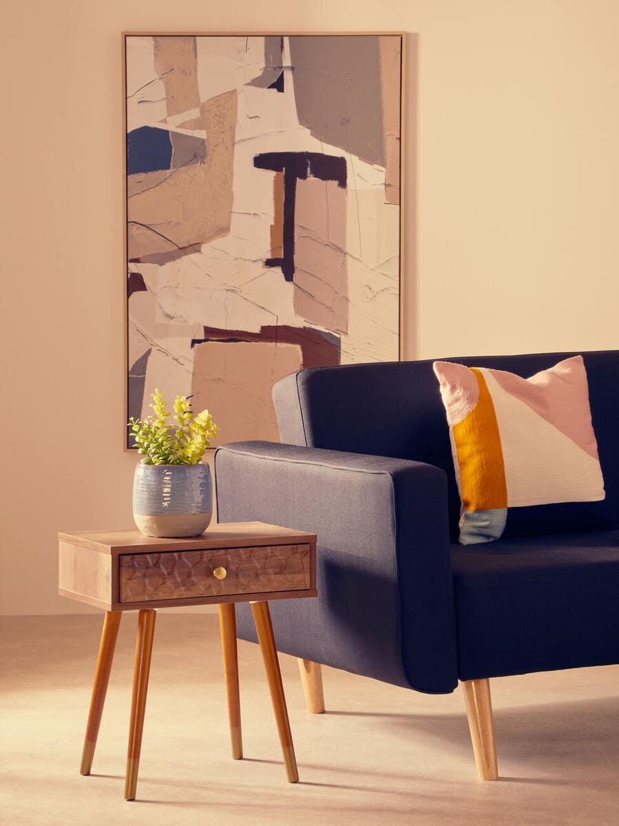 Wooden side table with drawer and plant beside navy fabric sofa and modern abstract art