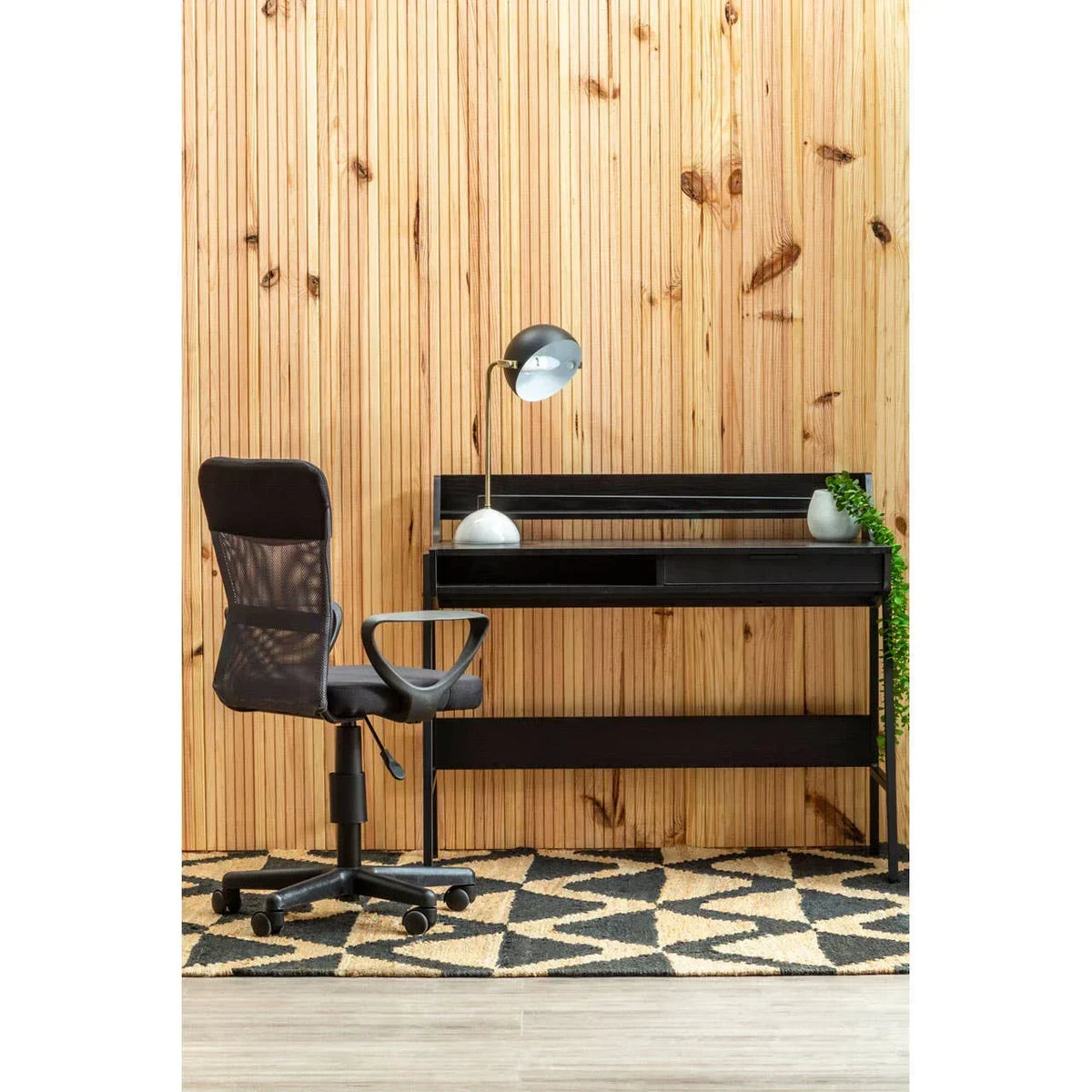 Black home office desk with mesh swivel chair, modern lamp, and plant against wood panel wall