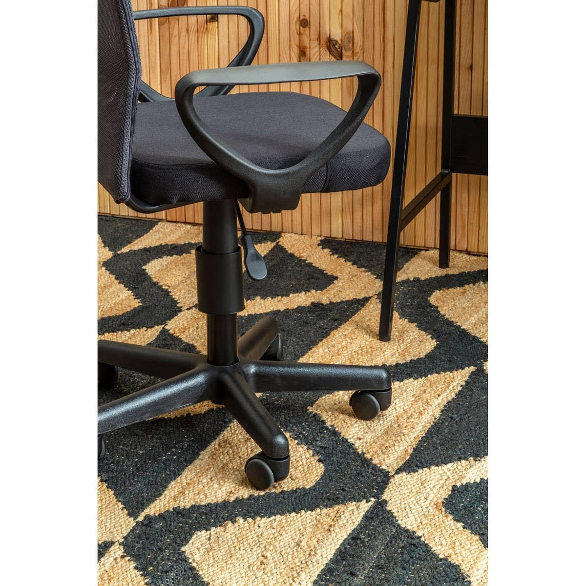 Black office chair with armrests on patterned rug beside wooden wall and desk