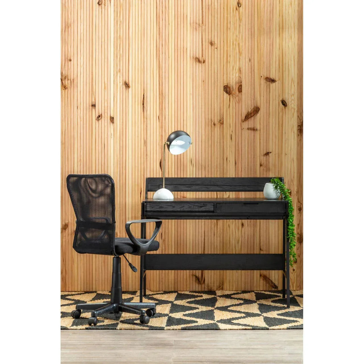 Black office desk and mesh chair set on patterned rug with table lamp and planter