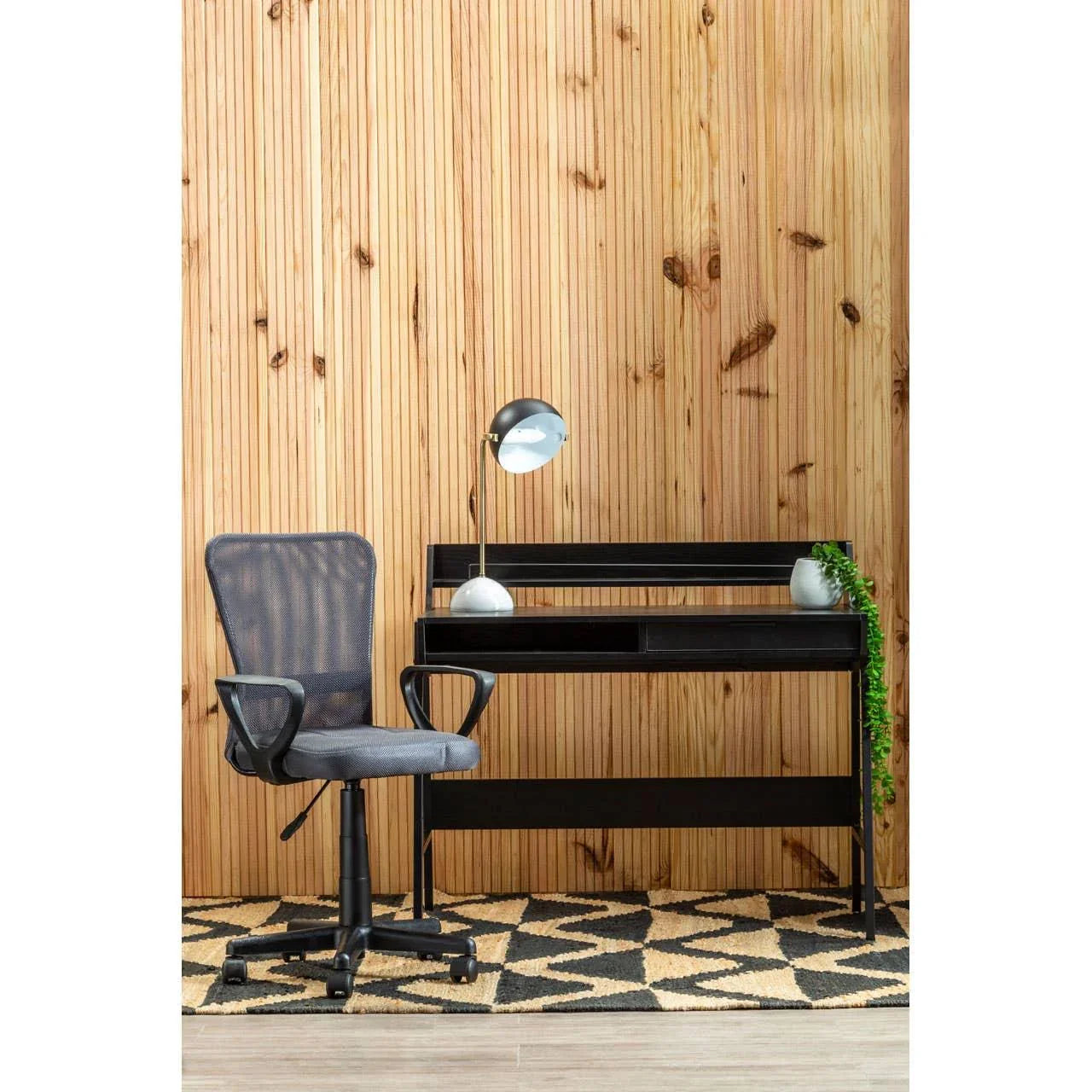 Black home office desk with mesh swivel chair, desk lamp, and plant against wood wall