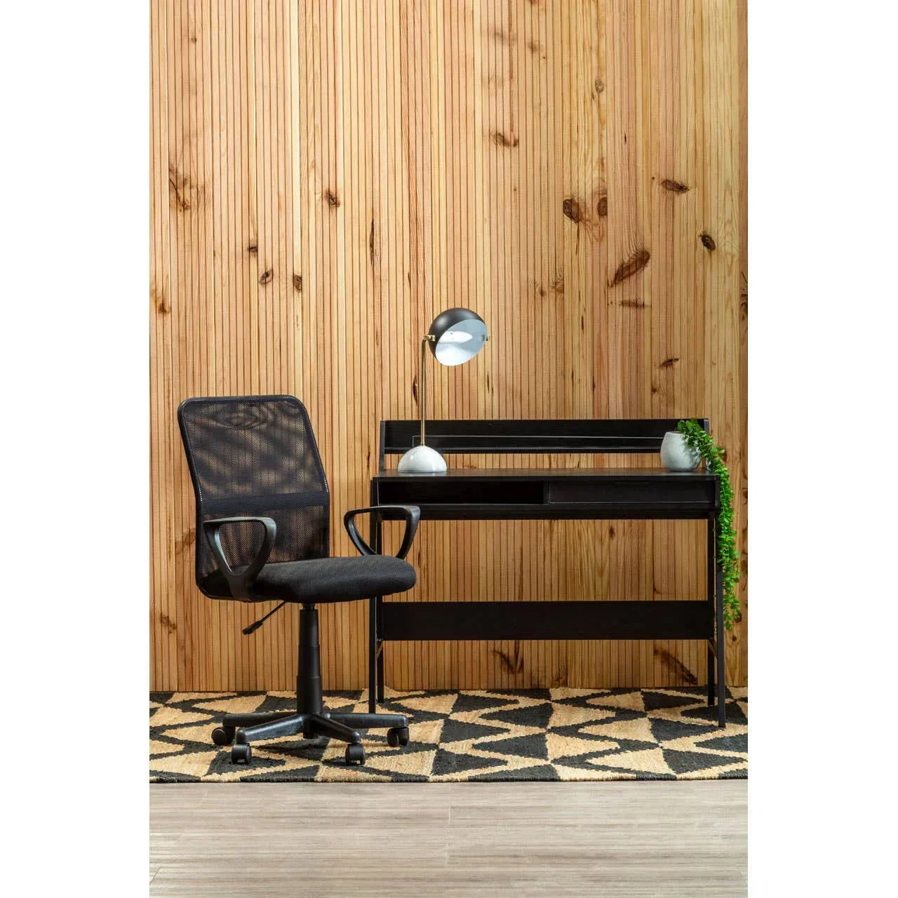 Black mesh office chair with black desk, desk lamp, and plant against wooden wall