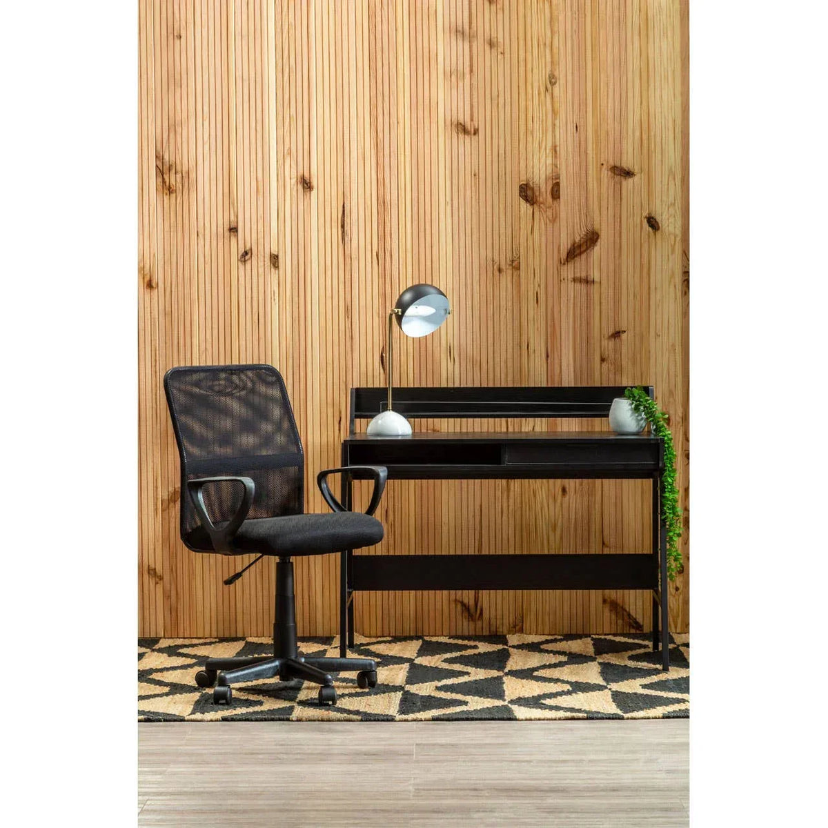 Black mesh office chair with black desk, desk lamp, and plant against wooden wall