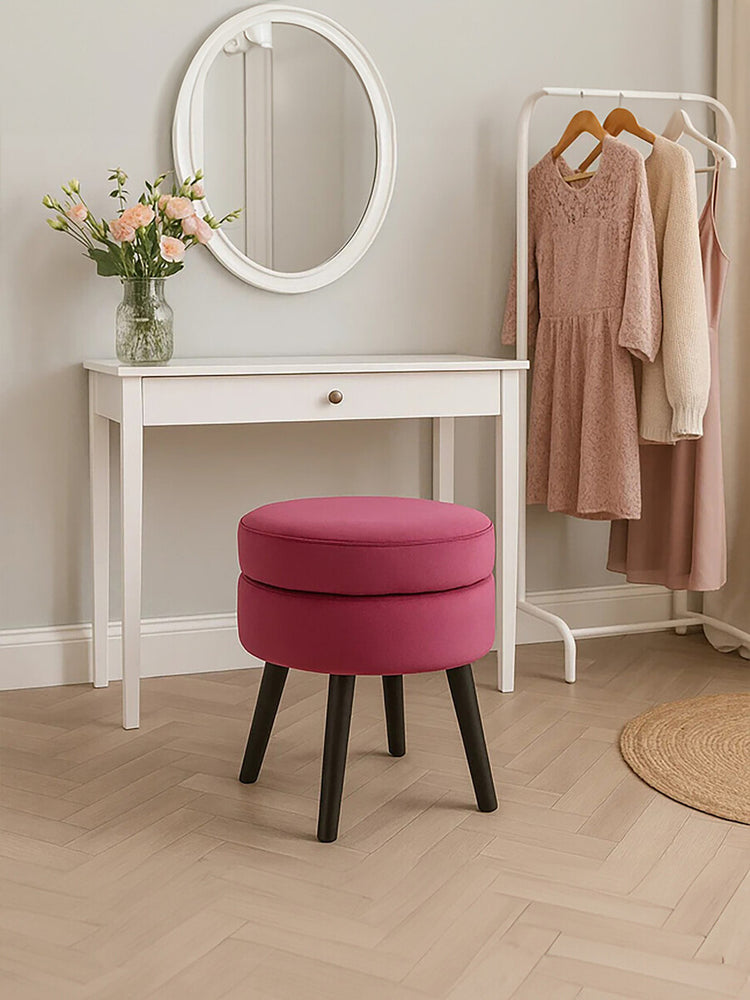 Round wine-colored velvet stool with a cushioned top and black wooden legs, positioned next to a white vanity table with a round mirror and a vase of flowers, set in a softly lit room with beige flooring