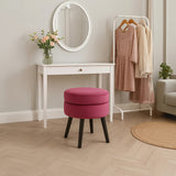 A round, wine-colored velvet stool with a plush top and black wooden legs is positioned in a bright room. Behind it, a white vanity table with a round mirror reflects soft lighting. A bouquet of flowers sits on the table, and two light-colored dresses hang on a rack nearby, adding a touch of elegance to the space. The floor features a light wood pattern, enhancing the cozy atmosphere