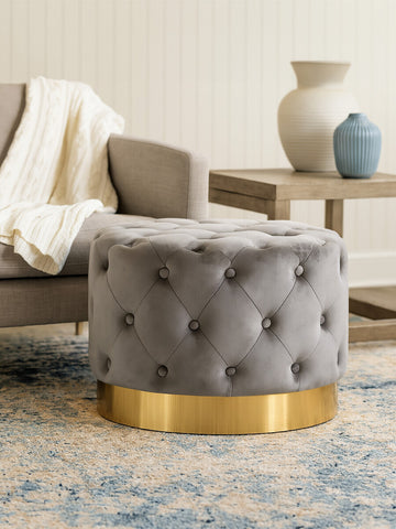 Grey velvet stool featuring a tufted design and a matte gold finish metal base, positioned next to a light-colored sofa with a throw blanket draped over it. A decorative vase and a small table are visible in the background, along with a patterned area rug