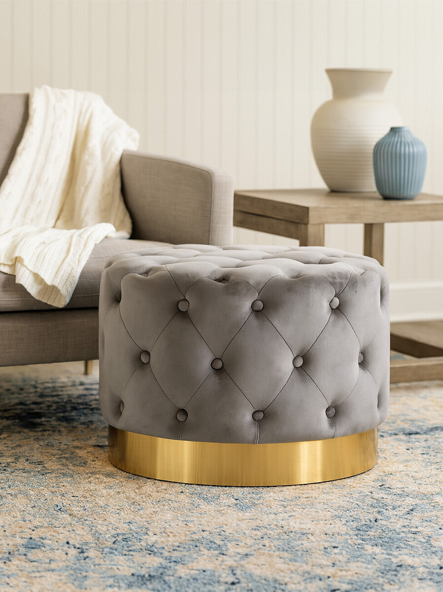 Grey velvet stool featuring a tufted design and a matte gold finish metal base, positioned next to a light-colored sofa with a throw blanket draped over it. A decorative vase and a small table are visible in the background, along with a patterned area rug