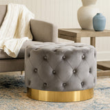 Grey velvet stool featuring a tufted design and a matte gold finish metal base, positioned next to a light-colored armchair and a wooden side table, with a decorative vase in the background