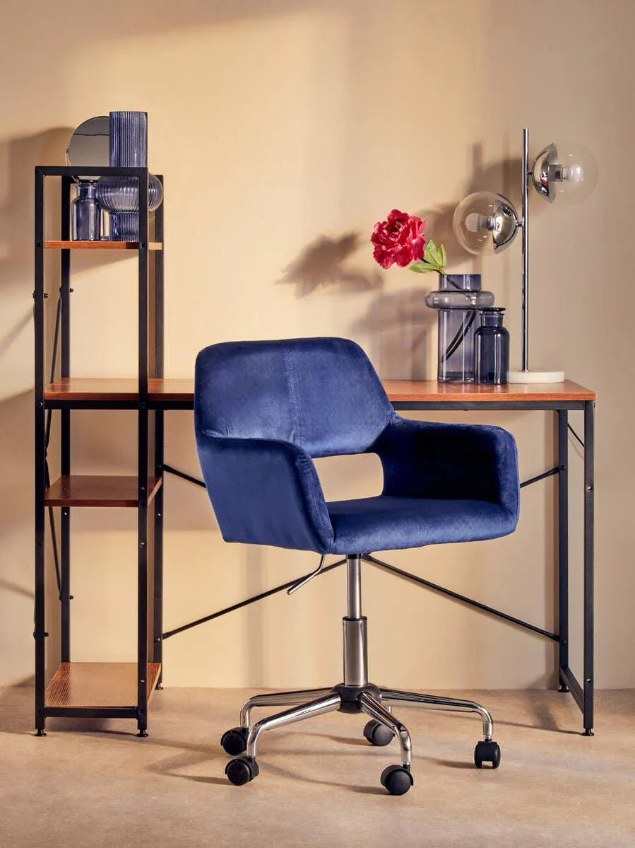 Blue velvet office chair with chrome base in front of wooden desk and black shelving unit