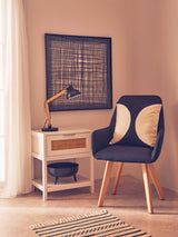White wood side table with one drawer and rattan detailing, positioned next to a modern chair with a patterned cushion. A small lamp is placed on the table, and a textured rug is visible on the floor. The background features a framed wall art piece and soft natural light coming through a window