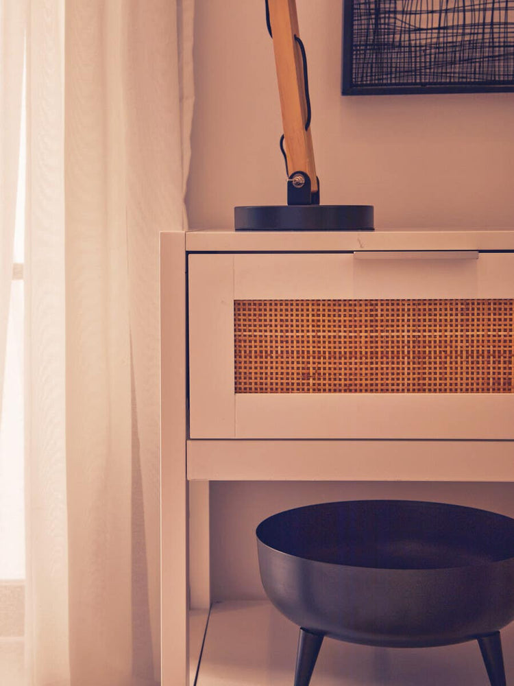 White wood side table with a rattan drawer front, featuring a single drawer and a flat top. A black lamp is placed on top, and a round black bowl sits on the floor beside the table. Soft natural light filters through sheer curtains in the background