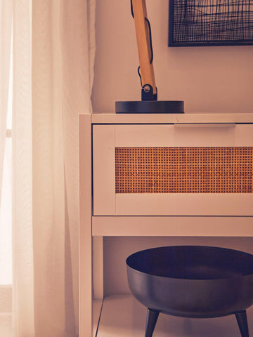 White wood side table with a rattan drawer front, featuring a single drawer and a flat top. A black lamp is placed on top, and a round black bowl sits on the floor beside the table. Soft natural light filters through sheer curtains in the background