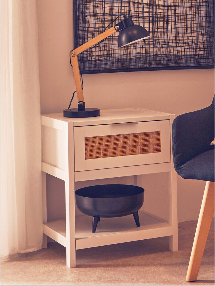 White wood side table featuring a rattan drawer front, a single drawer, and a lower shelf. A black lamp with a wooden arm is positioned on top, alongside a round black bowl. The table is set against a light-colored wall with a patterned artwork above it and a dark chair nearby. Natural light filters through sheer curtains