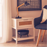 A white wood side table with a rattan drawer front, featuring one drawer and an open shelf below. A black bowl sits on the shelf, and a stylish black lamp with a yellow arm is positioned on top. A gray chair is partially visible next to the table, with soft curtains and a framed artwork in the background
