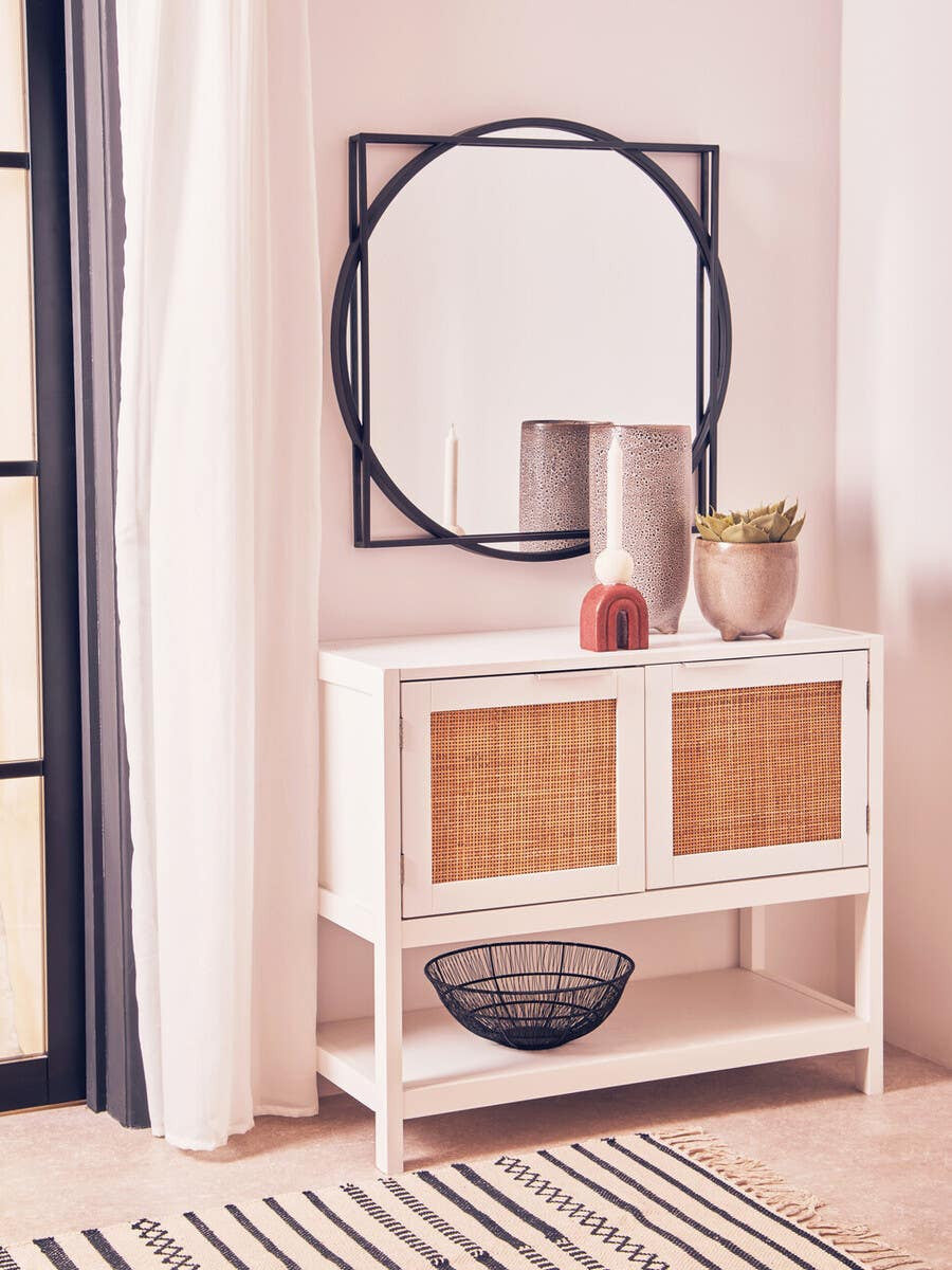 White wood side cabinet with two rattan drawers, placed against a light-colored wall. Above the cabinet, a round mirror with a decorative black frame reflects the room. A few decorative items, including a vase and a candle, are arranged on top of the cabinet, while a woven basket is visible in the open space below. A striped rug is partially visible on the floor