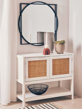 White wood side cabinet with two rattan-fronted drawers, placed beneath a round black-framed mirror. The cabinet is styled with decorative vases and a bowl on top, set against a light-colored wall and a striped rug