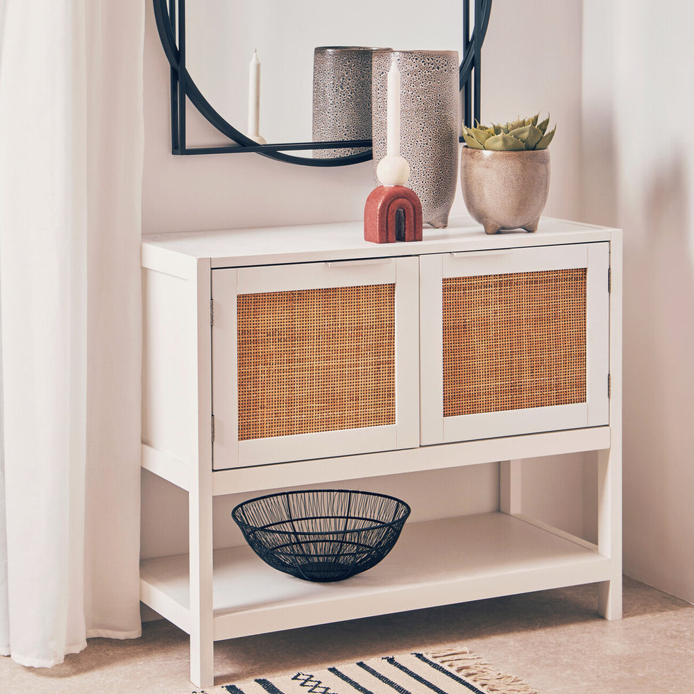 White wood side cabinet featuring two rattan-fronted drawers, a lower shelf, and decorative items on top, including vases and a bowl. A round mirror is mounted above the cabinet, reflecting the surrounding space