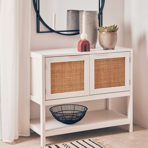 White wood side cabinet featuring two rattan-fronted drawers, a lower shelf, and decorative items on top, including vases and a bowl. A round mirror is mounted above the cabinet, reflecting the surrounding space