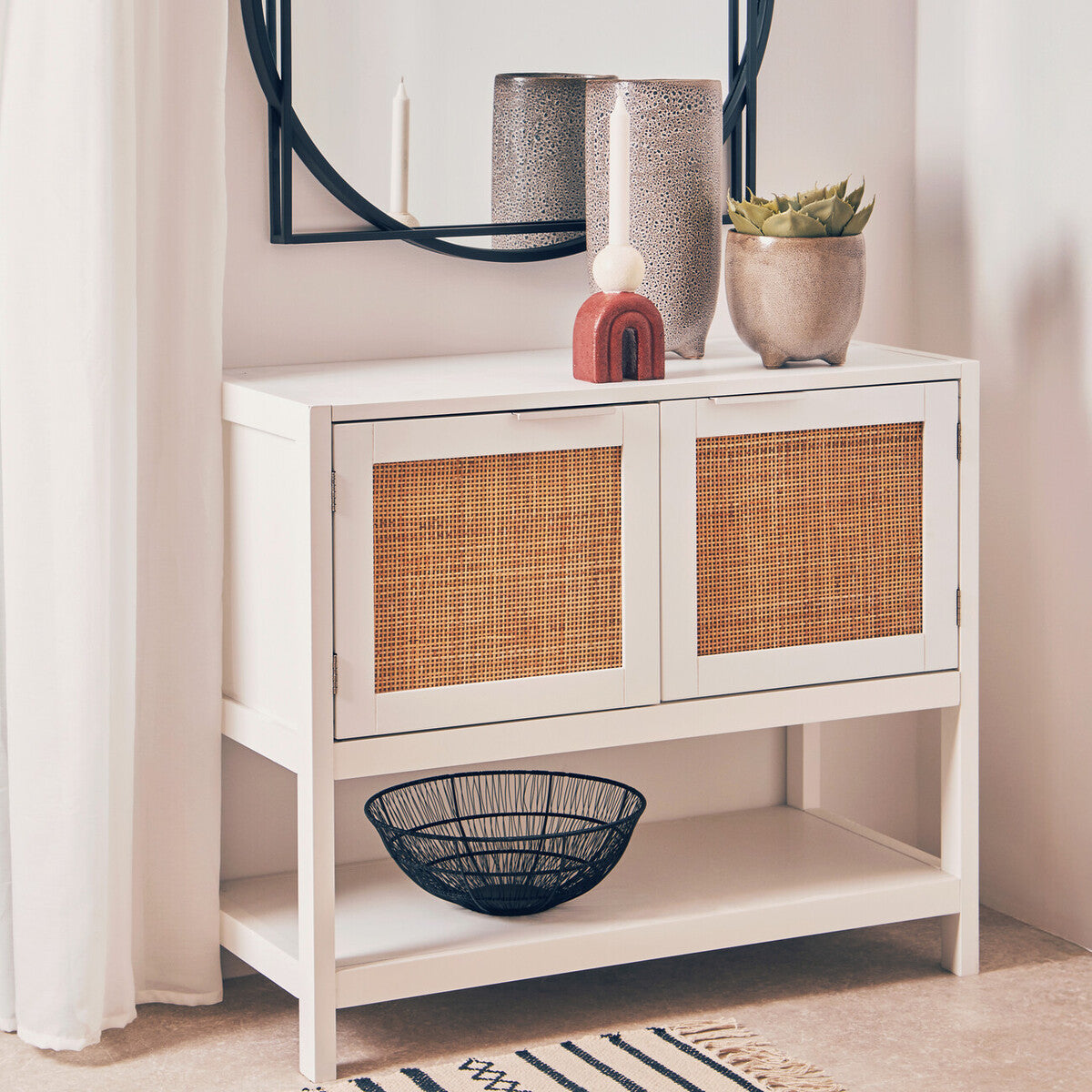 White wood side cabinet featuring two rattan-fronted drawers, a lower shelf, and decorative items on top, including vases and a bowl. A round mirror is mounted above the cabinet, reflecting the surrounding space