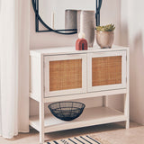 White wood side cabinet featuring two rattan-fronted drawers, a lower shelf, and decorative items on top, including vases and a bowl. A round mirror is mounted above the cabinet, reflecting the surrounding space
