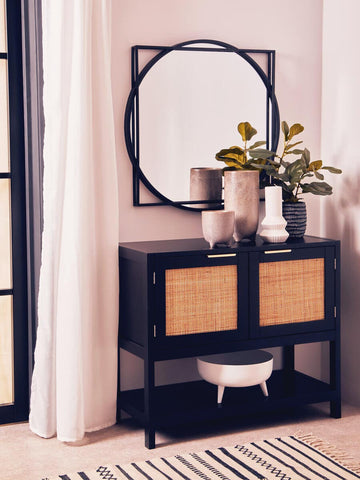 Black wood and rattan two-door side cabinet with a sleek design, featuring two rattan-woven doors and an open shelf below. A round mirror hangs above the cabinet, accompanied by decorative vases and greenery. The cabinet is positioned against a light-colored wall, with a patterned rug underneath