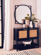 Black wood and rattan two-door side cabinet with a sleek design, featuring two rattan-woven doors and an open shelf below. A round mirror hangs above the cabinet, accompanied by decorative vases and greenery. The cabinet is positioned against a light-colored wall, with a patterned rug underneath
