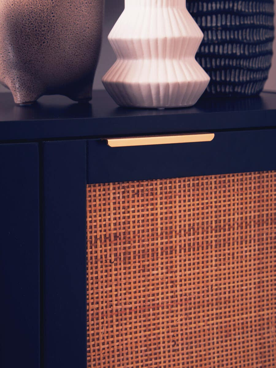 Black wood side cabinet featuring two doors with a rattan panel design. A sleek gold handle is positioned on the upper part of the cabinet. The cabinet is styled with decorative vases placed on top, showcasing a modern and elegant aesthetic