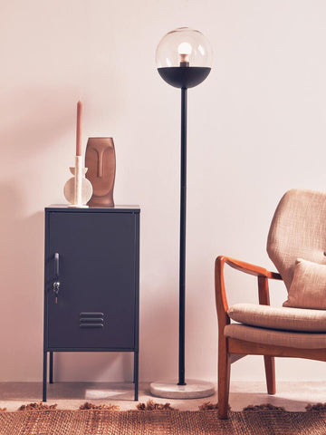 A grey one-door locker cabinet with a sleek design, positioned next to a modern floor lamp and a stylish armchair. The cabinet features a minimalist handle and is set against a light-colored wall, creating a contemporary living space ambiance