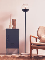 A grey one-door locker cabinet with a sleek design, positioned next to a modern floor lamp and a stylish armchair. The cabinet features a minimalist handle and is set against a light-colored wall, creating a contemporary living space ambiance