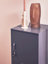 A grey one-door locker cabinet with a sleek design, featuring a lock and a smooth surface. On top of the cabinet, there is a tall, textured brown vase and a white candle holder with a candle, set against a soft pink wall