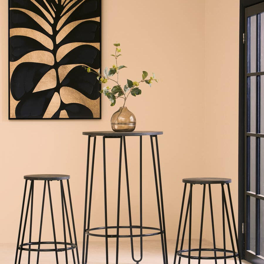 A modern three-piece bar table and stool set featuring a tall, round table with a sleek black metal frame and two matching stools. The table is adorned with a small vase containing greenery. Behind the set is a wall with a large, abstract black and gold artwork, and a window with black framing, allowing natural light to illuminate the scene. The background is a warm, neutral color