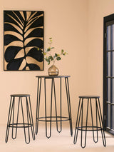 A modern three-piece bar table and stool set featuring a tall, round table with a sleek black metal frame and two matching stools. The table is adorned with a small vase containing greenery. Behind the set is a wall with a large, abstract black and gold artwork, and a window with black framing, allowing natural light to illuminate the scene. The background is a warm, neutral color